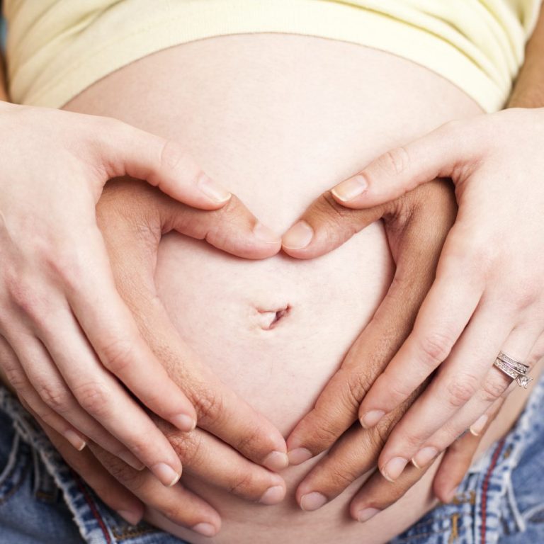 Hände formen ein Herz um einen schwangeren Bauch.